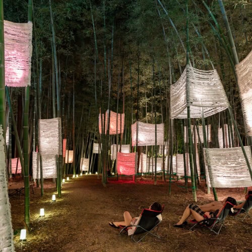 夜の竹林で、人物が寝椅子でくつろいでいます。頭上には、白く光る提灯のようなアート作品が吊るされ、道には小さなライトが並び、神秘的な雰囲気を醸し出しています。