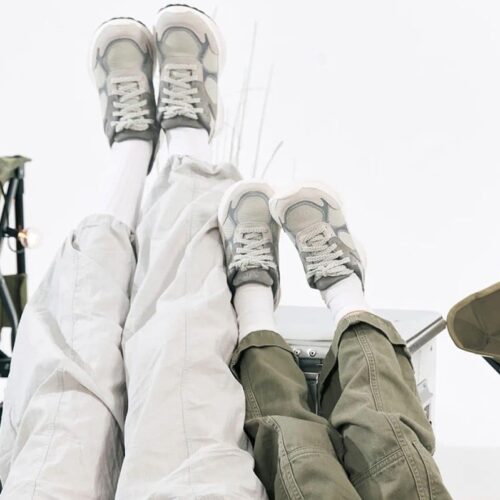 仰向けに寝て足を上げた2人。白いロングソックスとグレーのスポーティなスニーカーを履いている。カーキとライトグレーのカジュアルなパンツを履き、周囲にはキャンプ用のワゴンや椅子が見える。