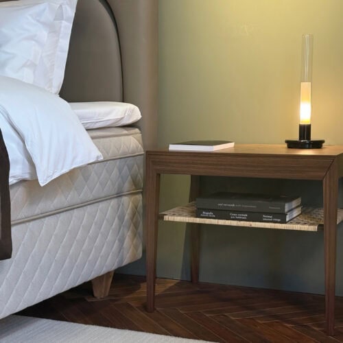 北欧デザインのサイドテーブルとベッドサイド。木製のナイトテーブルに籐(ラタン)の棚板、モダンなガラス製テーブルランプ。キルティングのベッドフレームが並ぶ寝室のディテール。