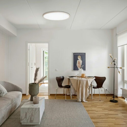 明るい自然光が差し込む、白を基調としたモダンな北欧風リビング・ダイニング。天井中央には白色に点灯する薄型の円形シーリングライトが設置されており、部屋全体を均一に照らしている。