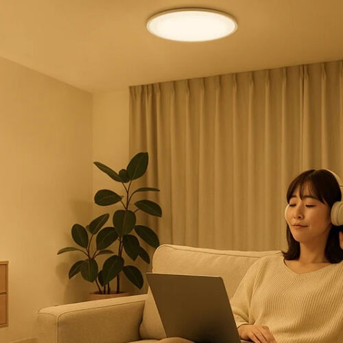 夜間、温かみのある電球色(暖色)に包まれた落ち着いたリビング。女性がソファでくつろぎながら、ヘッドホンで音楽を聴き、膝の上のノートパソコンを操作している。